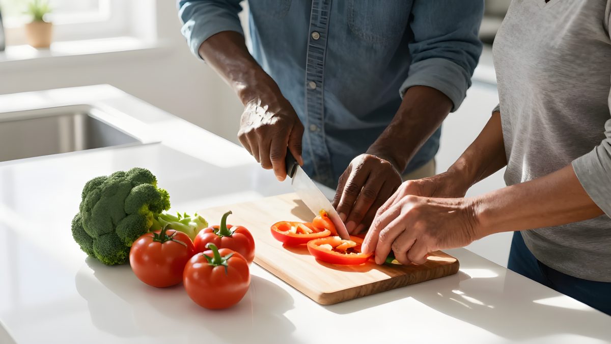 Två personer skär färska grönsaker tillsammans vid en vit köksbänk i solljus, med broccoli och tomater bredvid skärbrädan