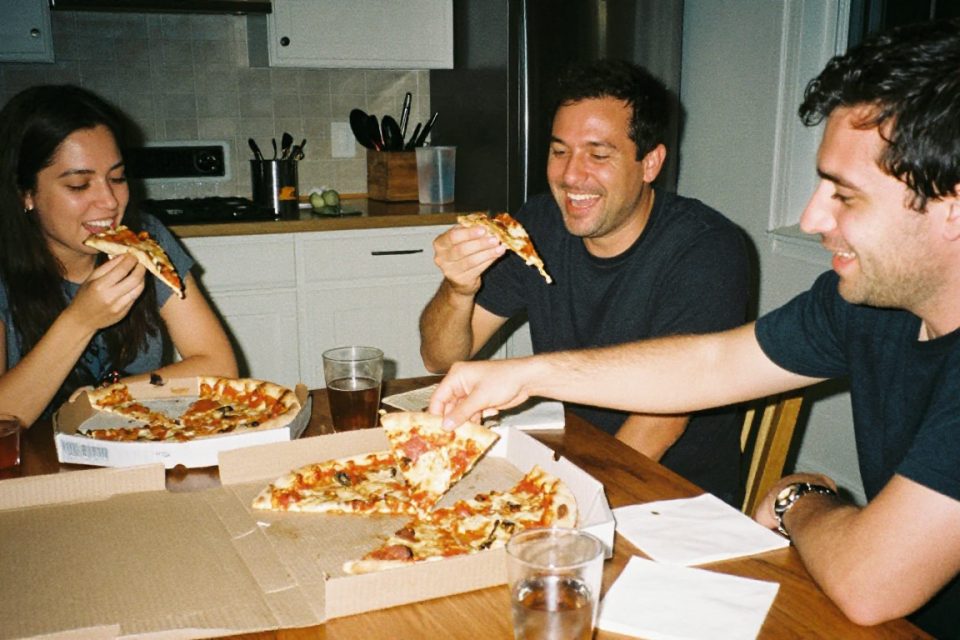 En man äter pizza direkt ur kartongen tillsammans med vänner vid ett köksbord en lördagskväll, en avslappnad fridag utan skuldkänslor.